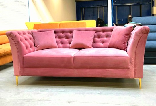 Różowa, welurowa sofa Chesterfield z trzema poduszkami, na złotych nóżkach, na tle innych sof w magazynie.