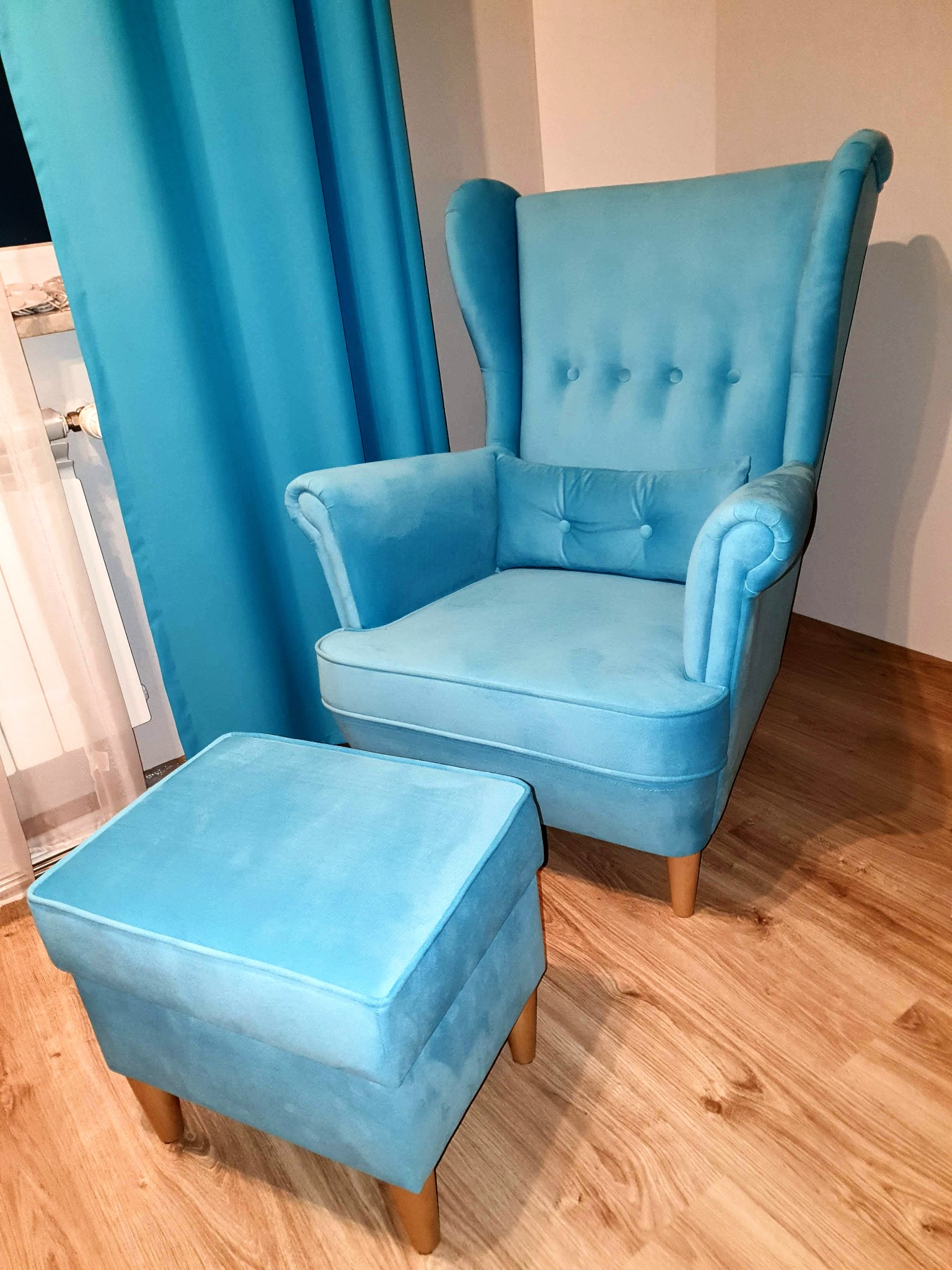 Turkusowy fotel uszak z podnóżkiem na drewnianej podłodze, obok okno z turkusową zasłoną.