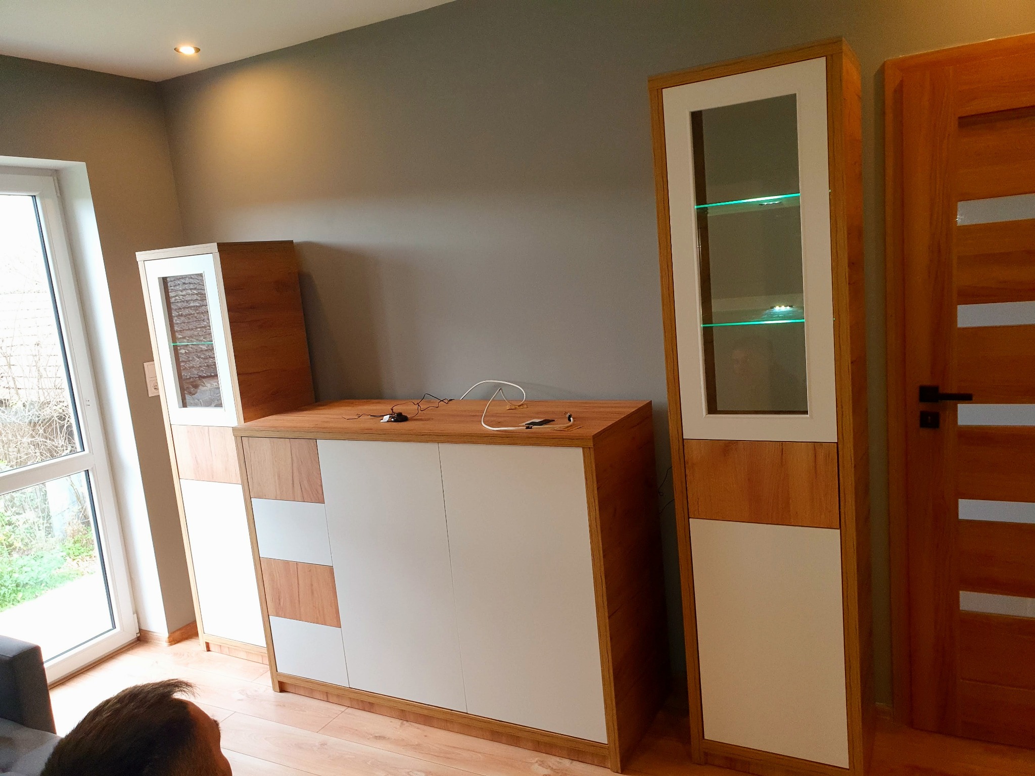 Zestaw mebli do salonu z drewna i białych frontów, w tym witryna z podświetlanymi półkami, komoda z szafkami i półkami oraz drzwi z drewnianymi panelami.