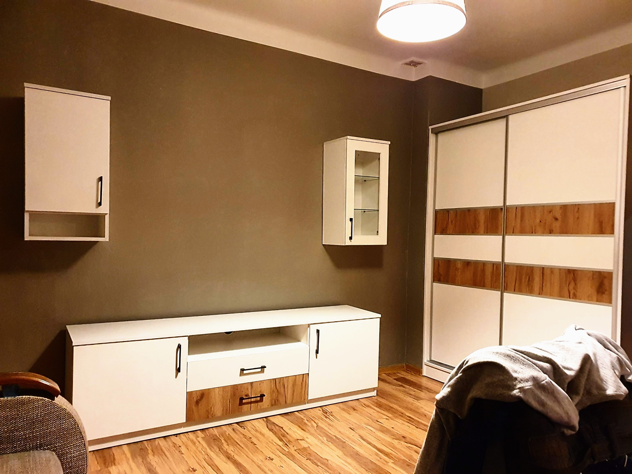 Nowoczesny salon z białymi meblami: szafa przesuwna z drewnianymi akcentami, szafki wiszące i szafka RTV, na podłodze panele w odcieniach drewna, ściana w kolorze taupe.