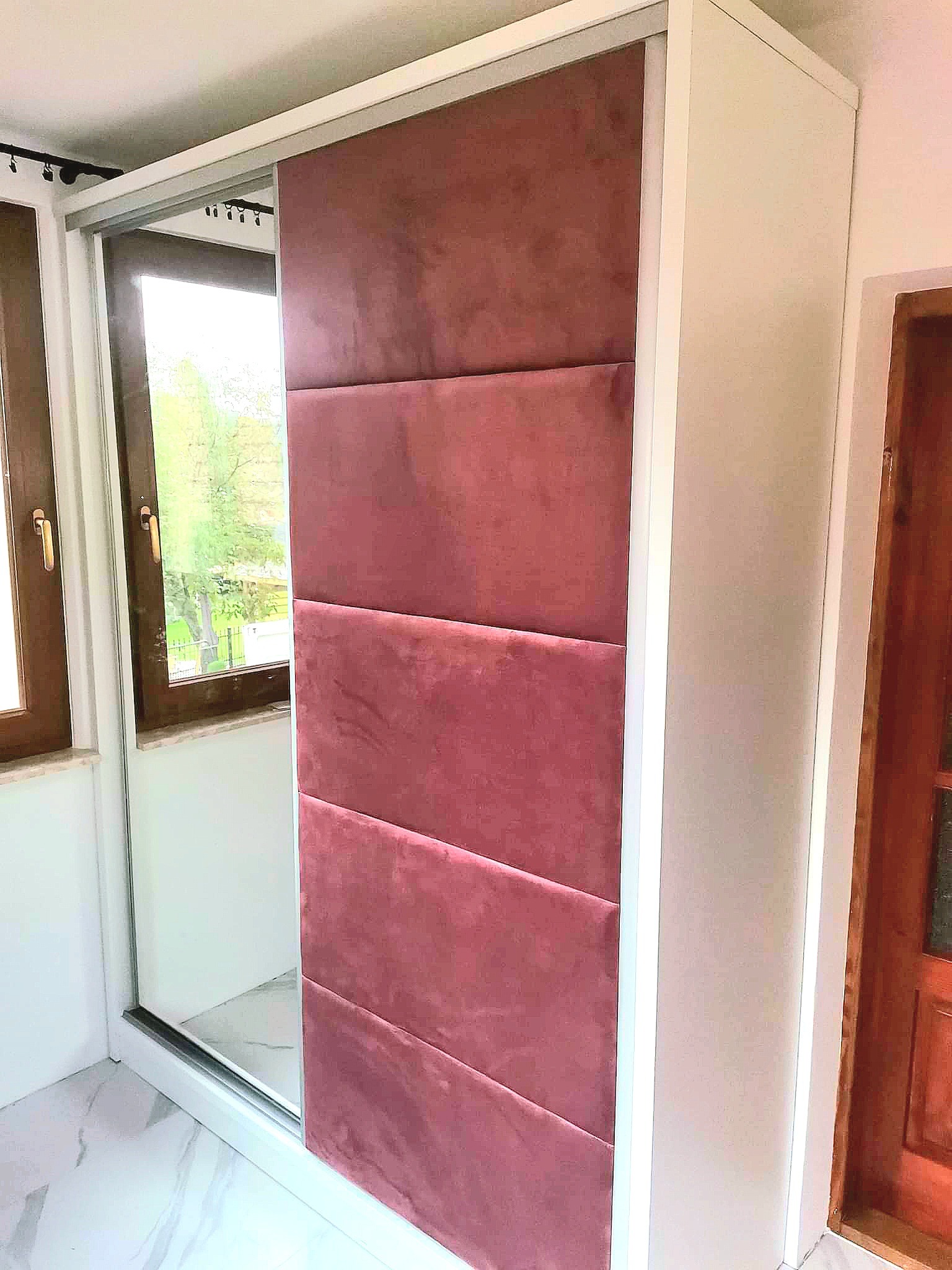 Biała szafa wnękowa z przesuwnymi drzwiami wykończonymi różowym, pikowanym materiałem, widoczna na tle okna.