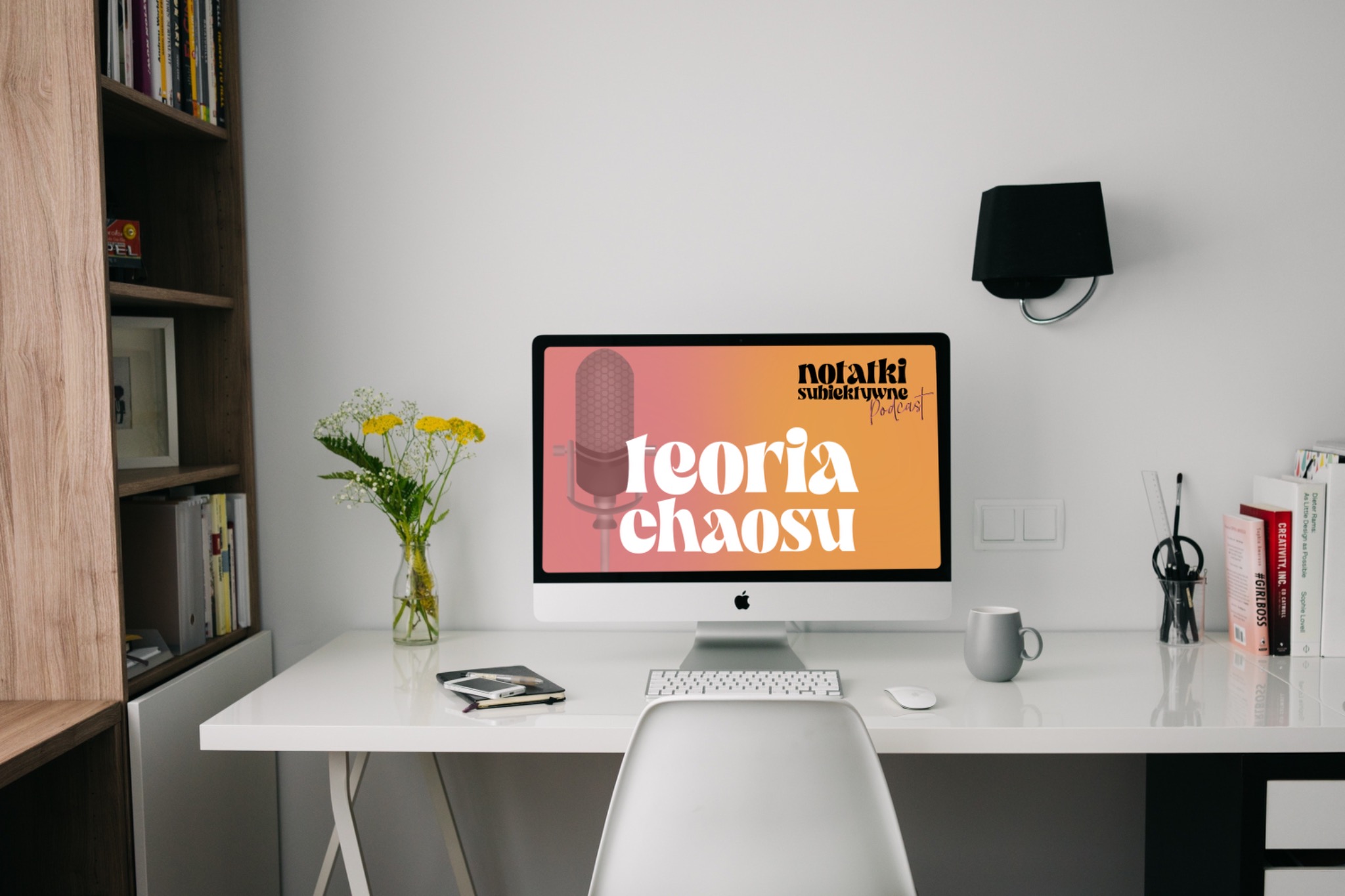 Białe biurko z komputerem iMac wyświetlającym grafikę podcastu 'Teoria chaosu', obok wazon z żółtymi kwiatami, szary kubek i przybornik na długopisy, w tle regał z książkami i lampa ścienna.