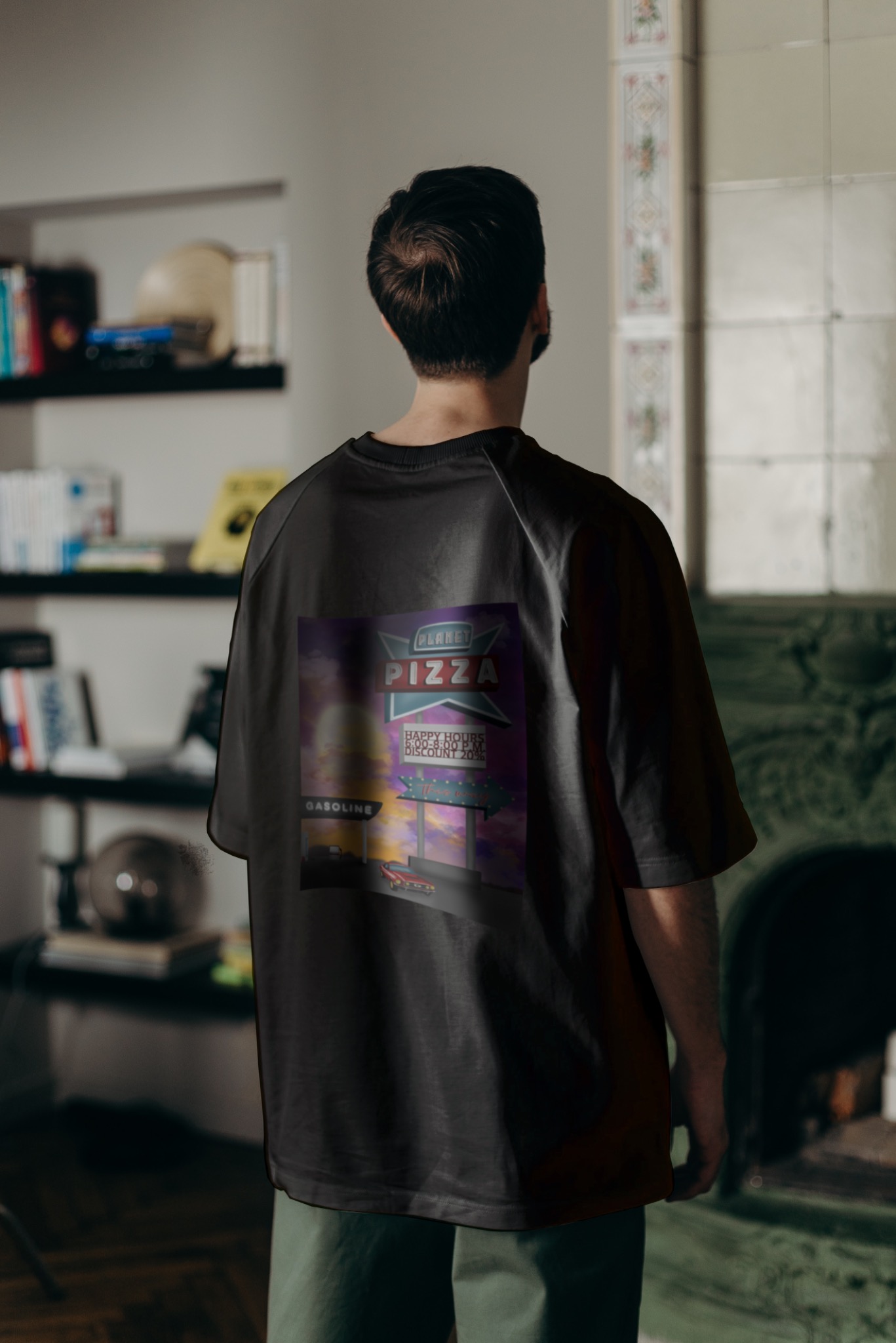 Mężczyzna tyłem w czarnej koszulce z nadrukiem retro reklamy pizzerii 'Planet Pizza' z napisem 'Happy Hours 6:00-8:00 PM, Discount 20%', stojący w pomieszczeniu z półkami na książki i dekoracyjnym...