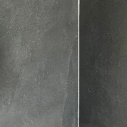 Szczeg&oacute;łowe ujęcie narożnika wyłożonego dużymi, ciemnoszarymi płytkami imitującymi kamień z cienkimi, jasnoszarymi fugami, z widocznym fragmentem drewnopodobnej płytki podłogowej.