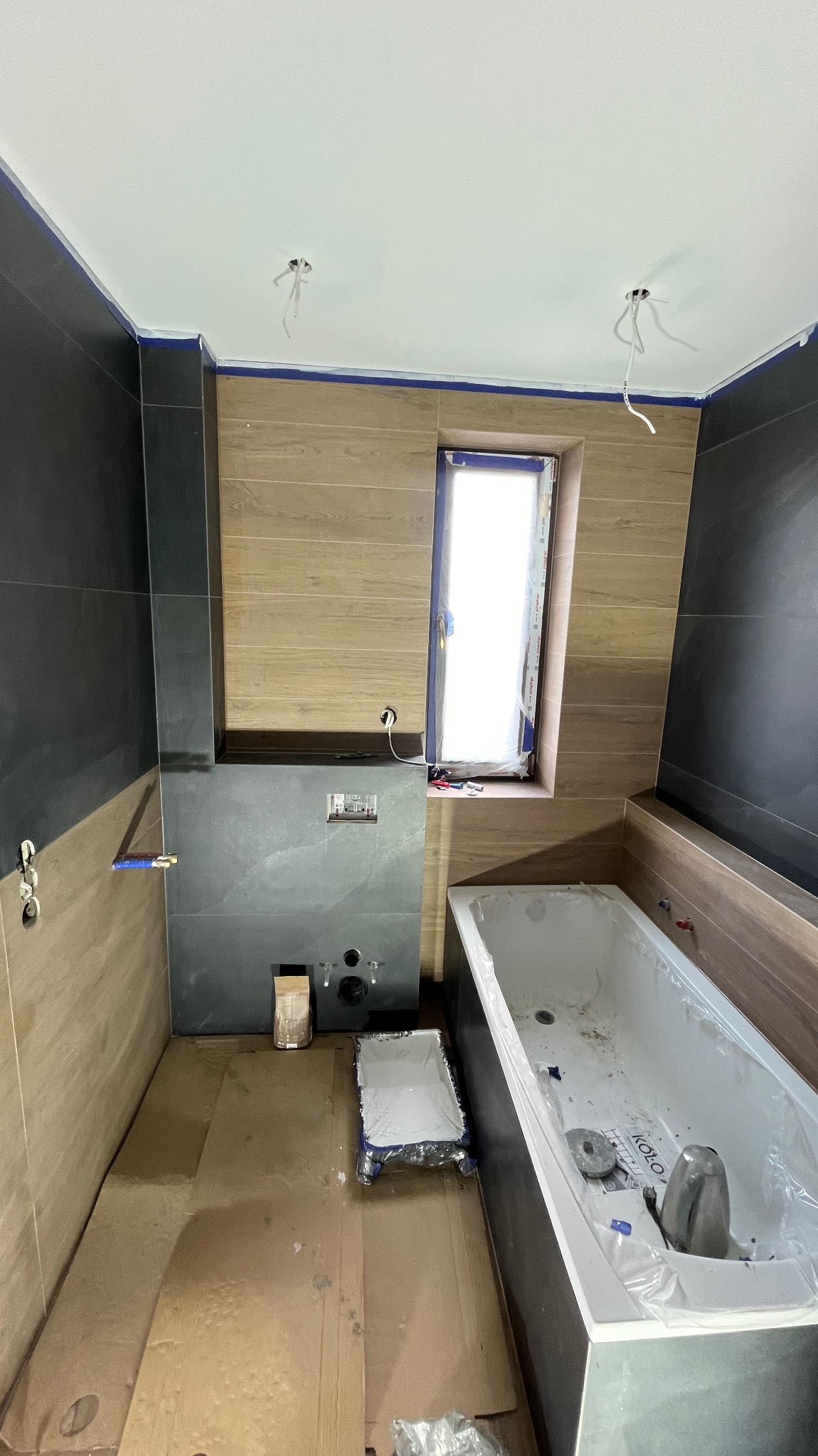 Łazienka w trakcie wykańczania: częściowo ułożone płytki imitujące drewno i grafitowe, wanna zabezpieczona folią, widoczne puszki elektryczne i przewody, kartony na podłodze.