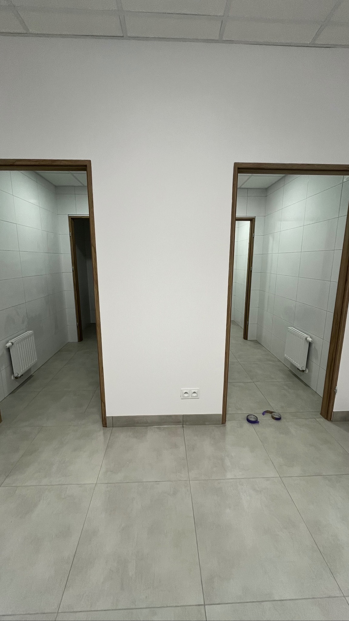 Dwa wejścia do łazienek z białymi płytkami na ścianach i grzejnikami, pomiędzy nimi fragment białej ściany z gniazdkiem elektrycznym, na podłodze leżą fioletowe klapki.