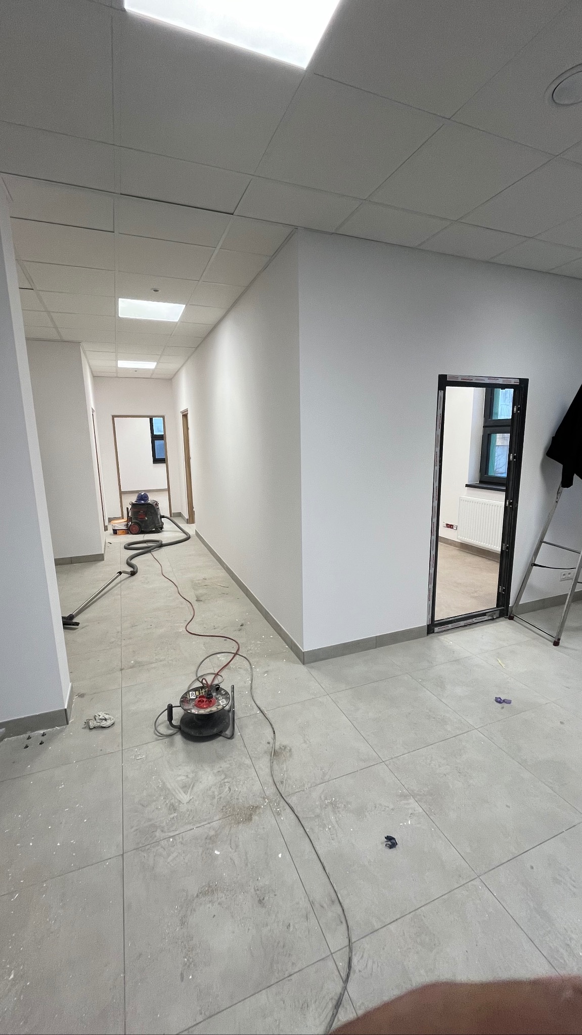 Korytarz w trakcie prac wykończeniowych, ściany świeżo pomalowane na biało, widoczny odkurzacz przemysłowy i zwinięty przedłużacz na podłodze z szarych płytek, ślady pyłu i zabrudzeń po remoncie.