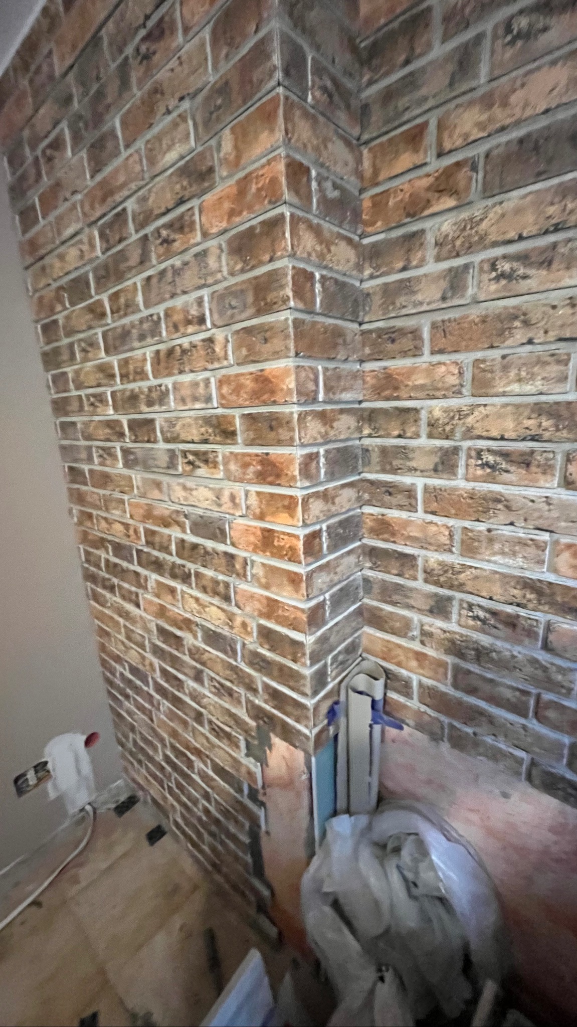 Fragment ściany wykończonej cegłą klinkierową w odcieniach brązu i beżu, widoczny narożnik, niedokończone prace, zabezpieczone rury i folia malarska na podłodze.