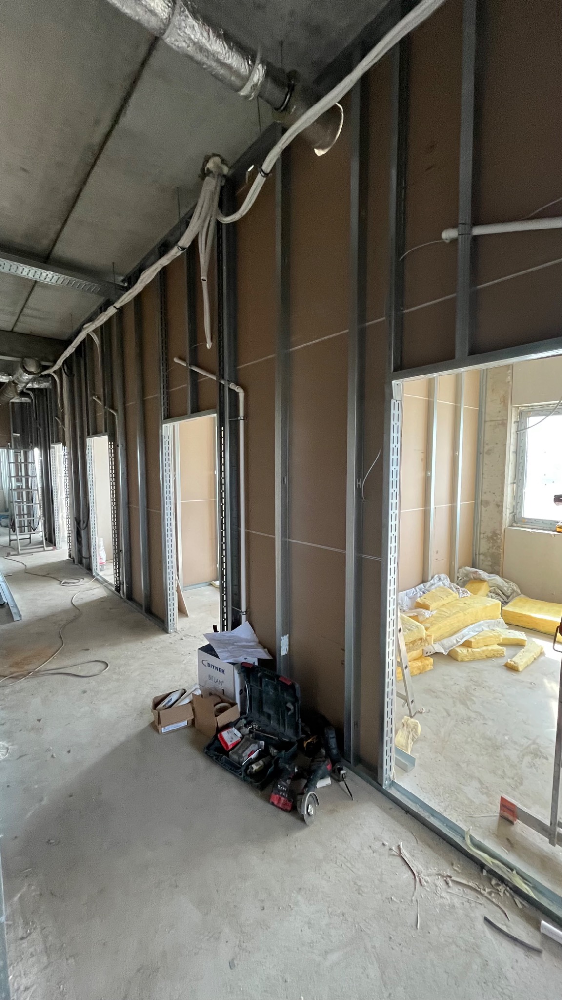 Montaż płyt karton-gips na metalowym stelażu w surowym wnętrzu. Widoczna izolacja, narzędzia i instalacje pod sufitem. Etap prac wykończeniowych.