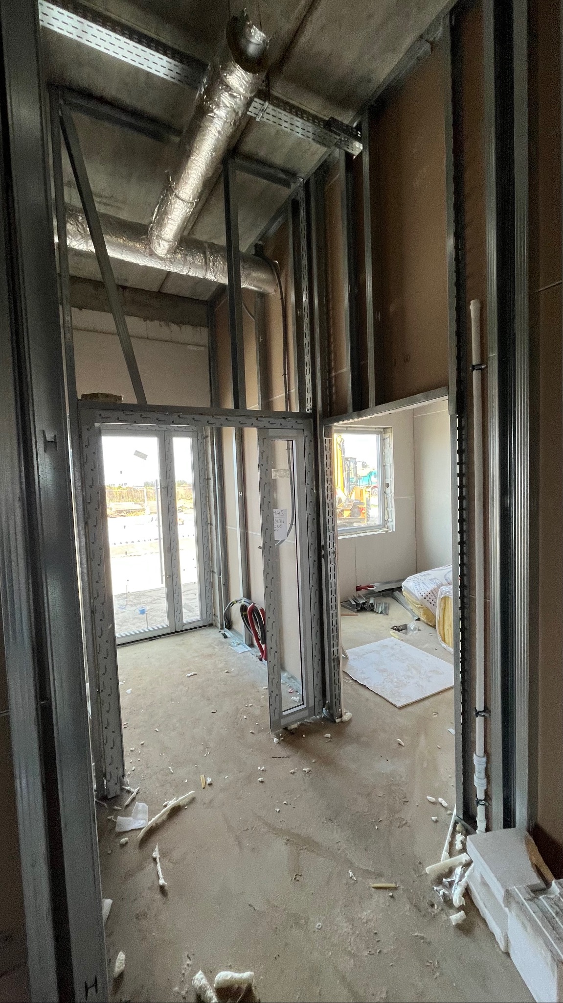Wnętrze w trakcie prac: metalowa konstrukcja ścian działowych, widoczne instalacje pod sufitem, okno z widokiem na budowę. Surowy stan wykończenia, podłoga z resztkami materiałów.