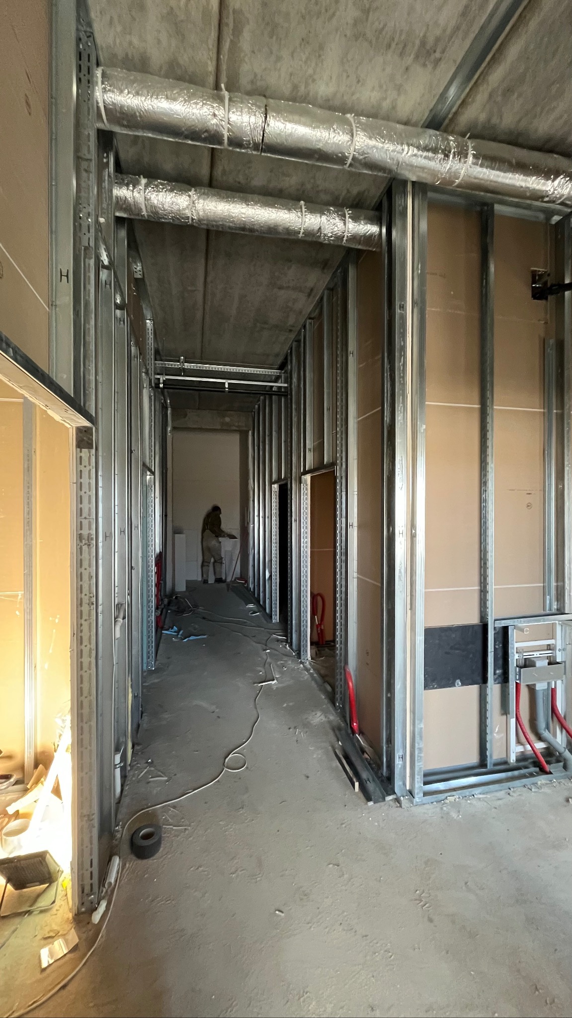 Montaż metalowej konstrukcji szkieletowej ścian działowych z płyt karton-gips w długim korytarzu. Widoczne instalacje i wentylacja pod stropem.