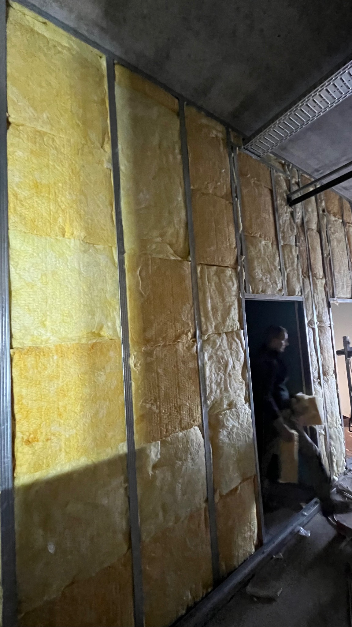 Ocieplanie ścian wełną mineralną w metalowej konstrukcji szkieletowej. Widoczna osoba w tle, w trakcie montażu izolacji. Surowy betonowy strop.