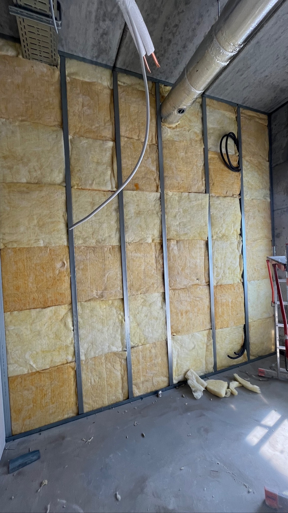 Ściana w trakcie ocieplania wełną mineralną, widoczne profile metalowe, rury wentylacyjne i kable. Fragment drabiny po prawej. Surowy stan wykończenia.