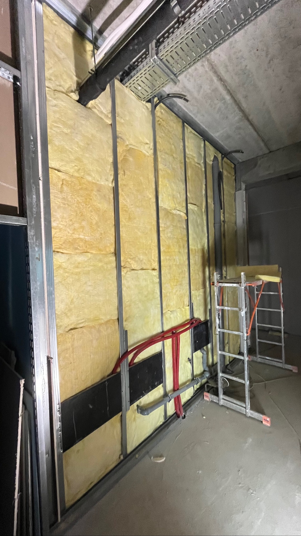Montaż izolacji z wełny mineralnej w metalowym stelażu ściany, widoczne rury instalacyjne i drabina. Prace wykończeniowe wnętrz w surowym stanie.
