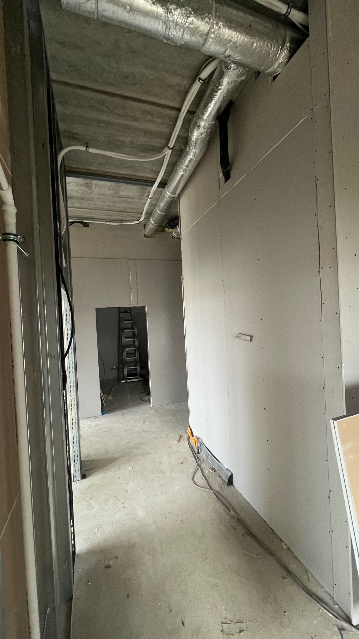 Wnętrze w trakcie prac: widoczne płyty karton-gips na ścianach, metalowe profile i instalacje pod sufitem. W tle drabina. Surowy stan wykończenia.