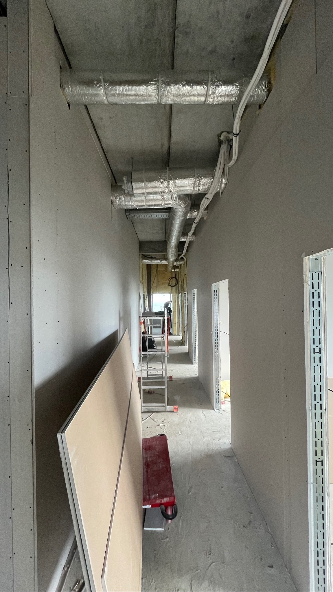 Korytarz w trakcie montażu płyt gipsowo-kartonowych na ścianach i suficie. Widoczne instalacje wentylacyjne i elektryczne pod sufitem. Na podłodze płyta i wózek transportowy.