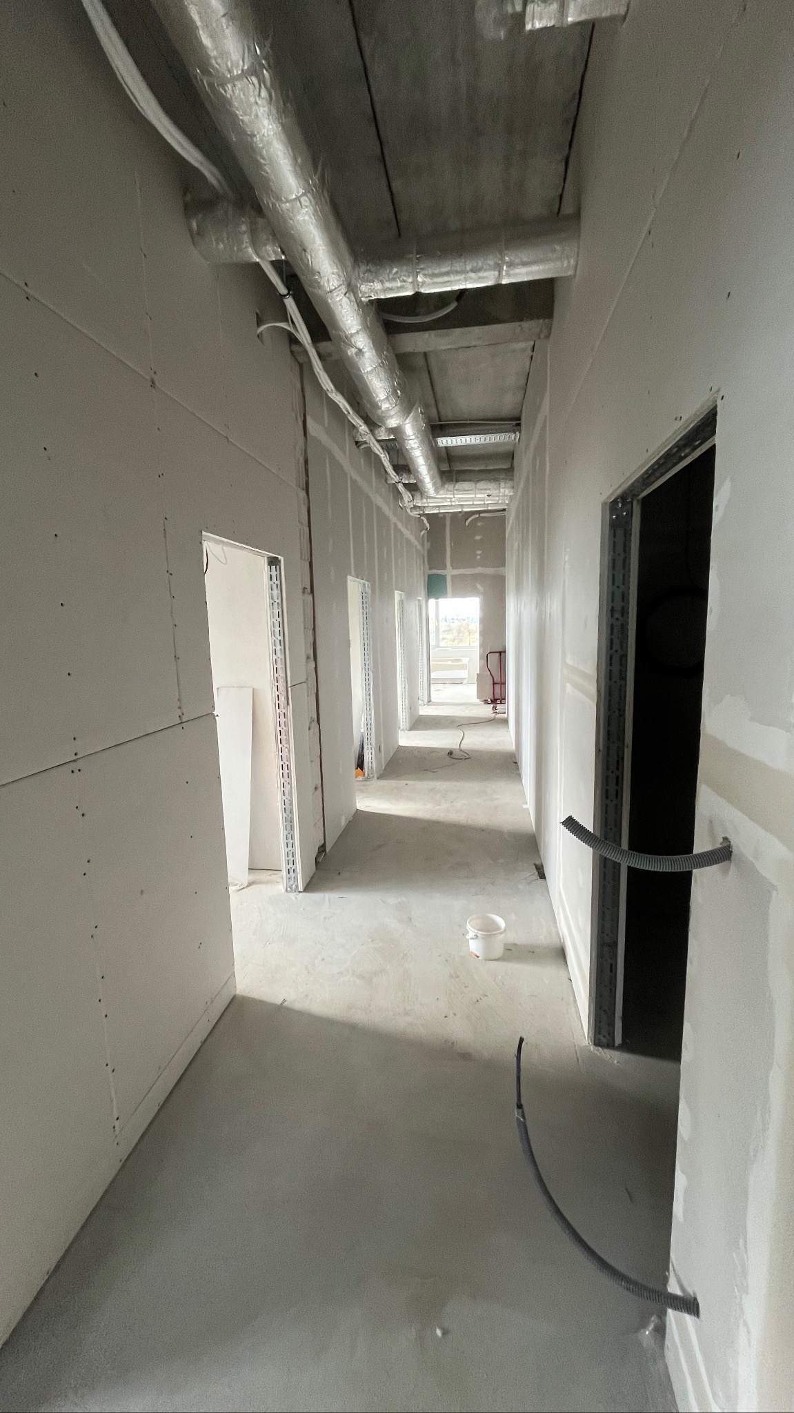 Korytarz w trakcie wykańczania: ściany z płyt gipsowo-kartonowych, widoczne instalacje pod sufitem, surowa podłoga. Białe ściany czekają na dalsze prace wykończeniowe.