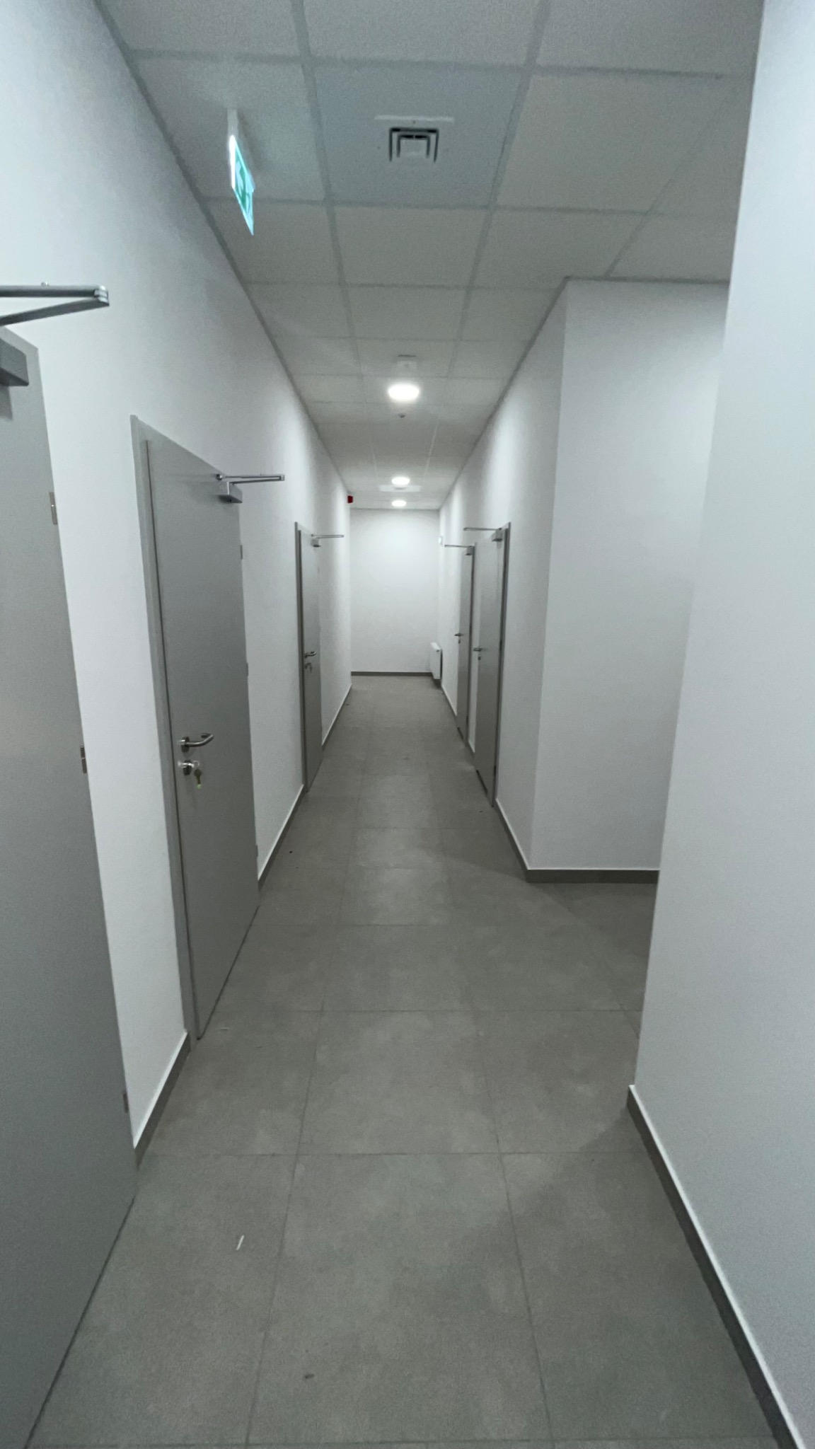 Wykończony korytarz biurowy w Pszczynie z szarymi drzwiami i płytkami podłogowymi. Widoczny znak ewakuacyjny i oświetlenie sufitowe.