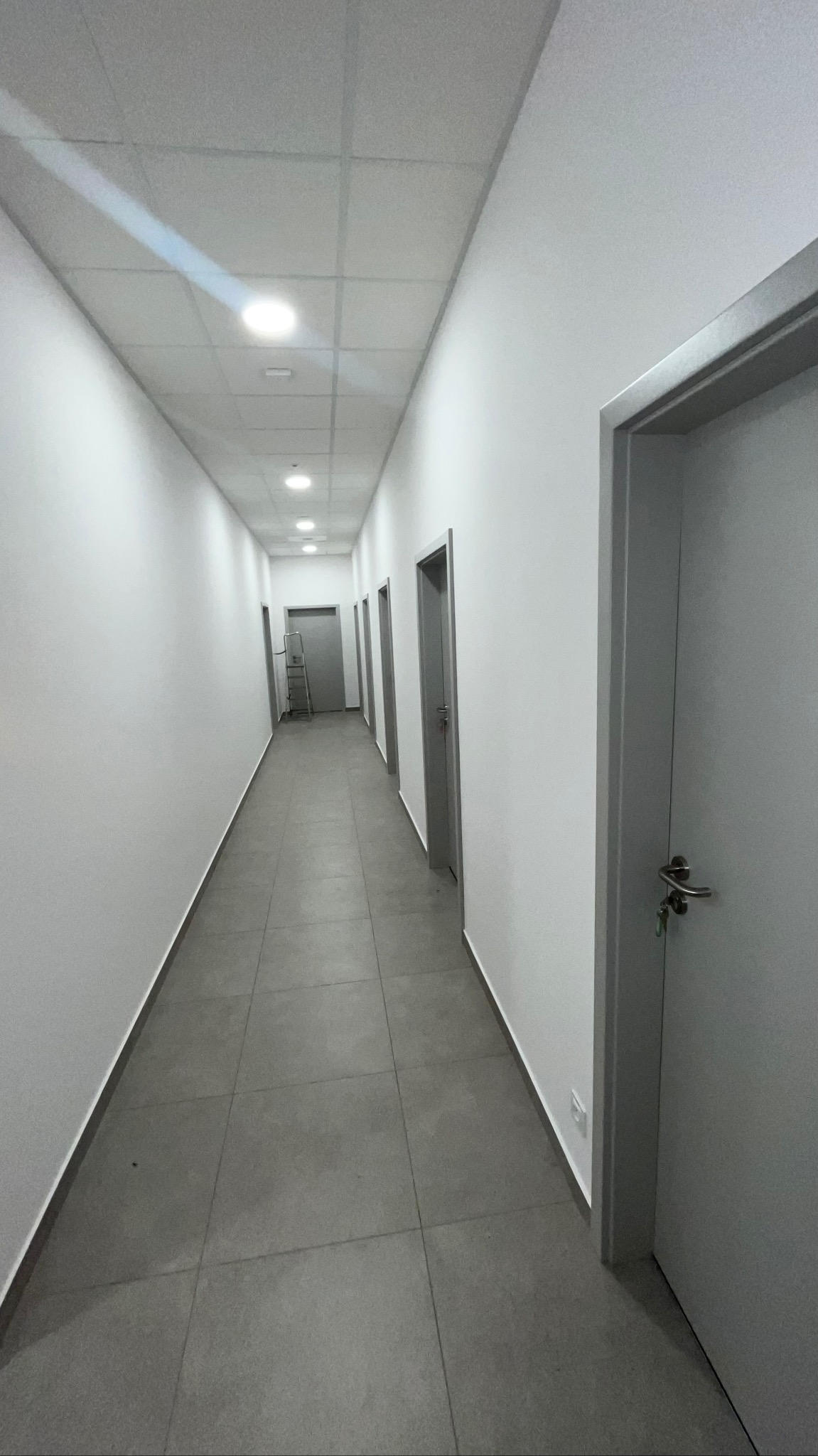 Długi, jasny korytarz z szarymi drzwiami i płytkami podłogowymi. W oddali widoczna drabina. Oświetlenie sufitowe równomiernie rozłożone.