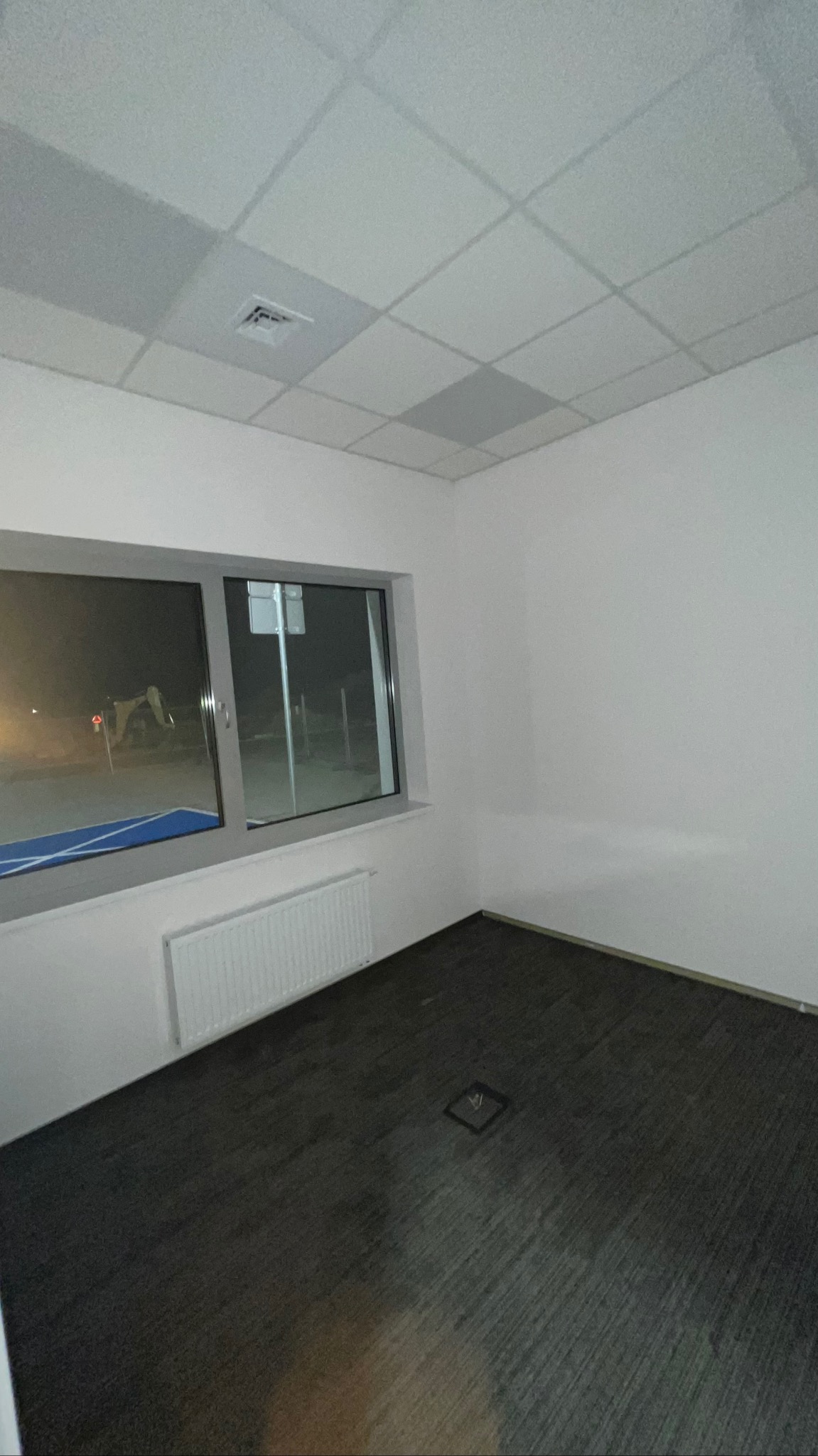 Wnętrze pokoju po remoncie: ciemna podłoga, białe ściany, sufit kasetonowy z kratką wentylacyjną, okno z widokiem na zewnątrz. Minimalistyczny design.