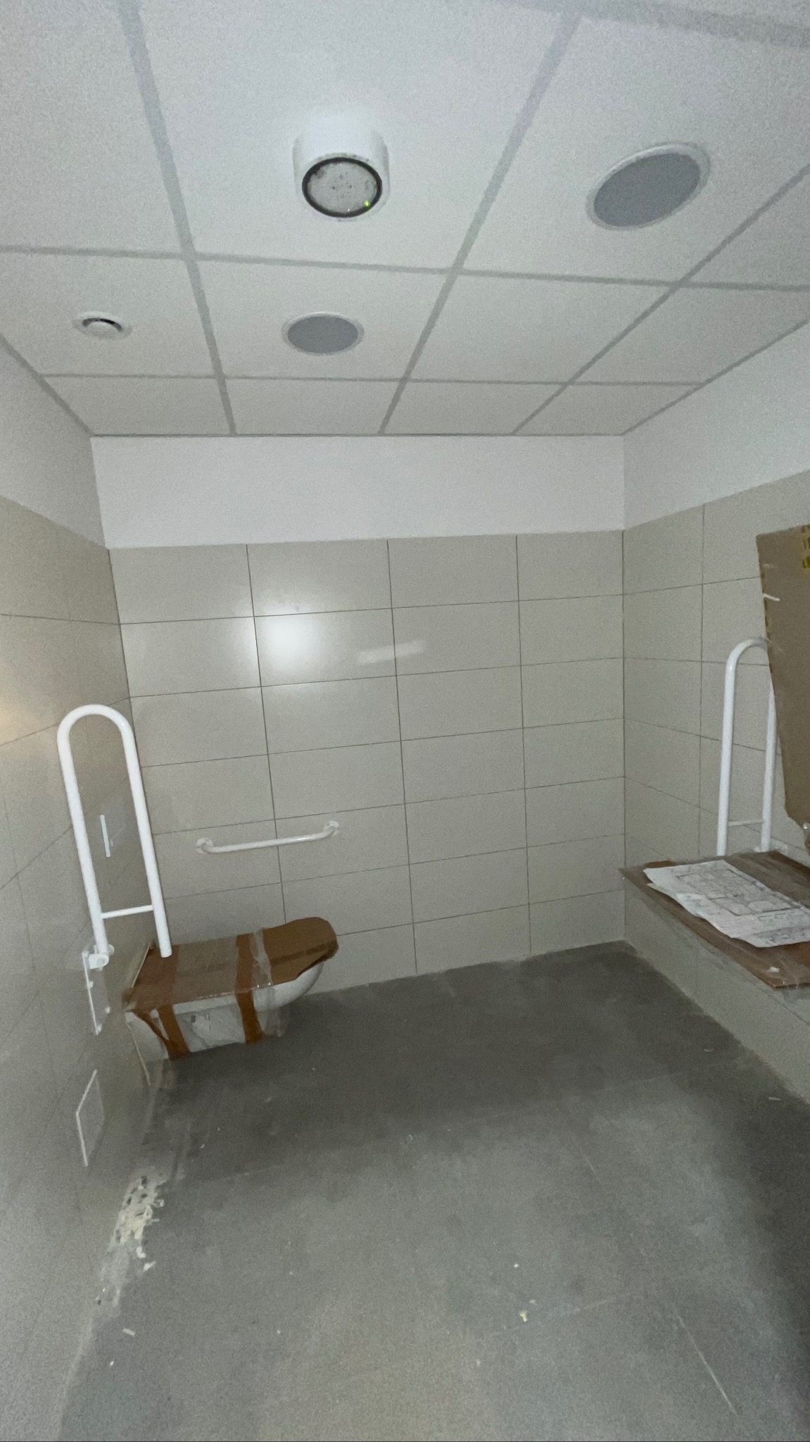 Wykończona łazienka z beżowymi płytkami, uchwytami dla niepełnosprawnych i zabezpieczoną toaletą. Widoczne oświetlenie sufitowe i szara podłoga.