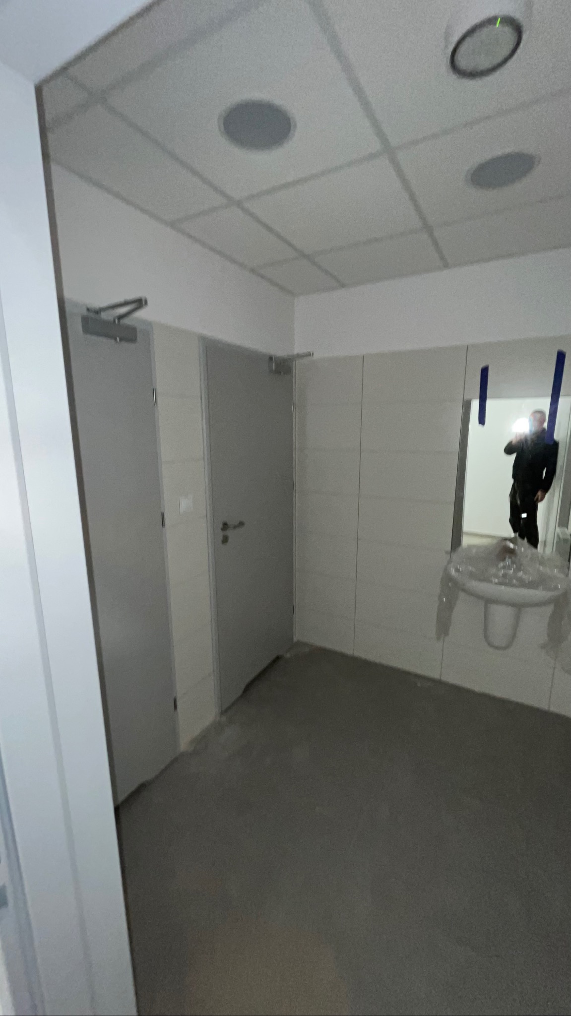 Wnętrze łazienki po remoncie z dwiema szarymi drzwiami, białą umywalką zabezpieczoną folią i lustrem, w którym odbija się osoba robiąca zdjęcie.