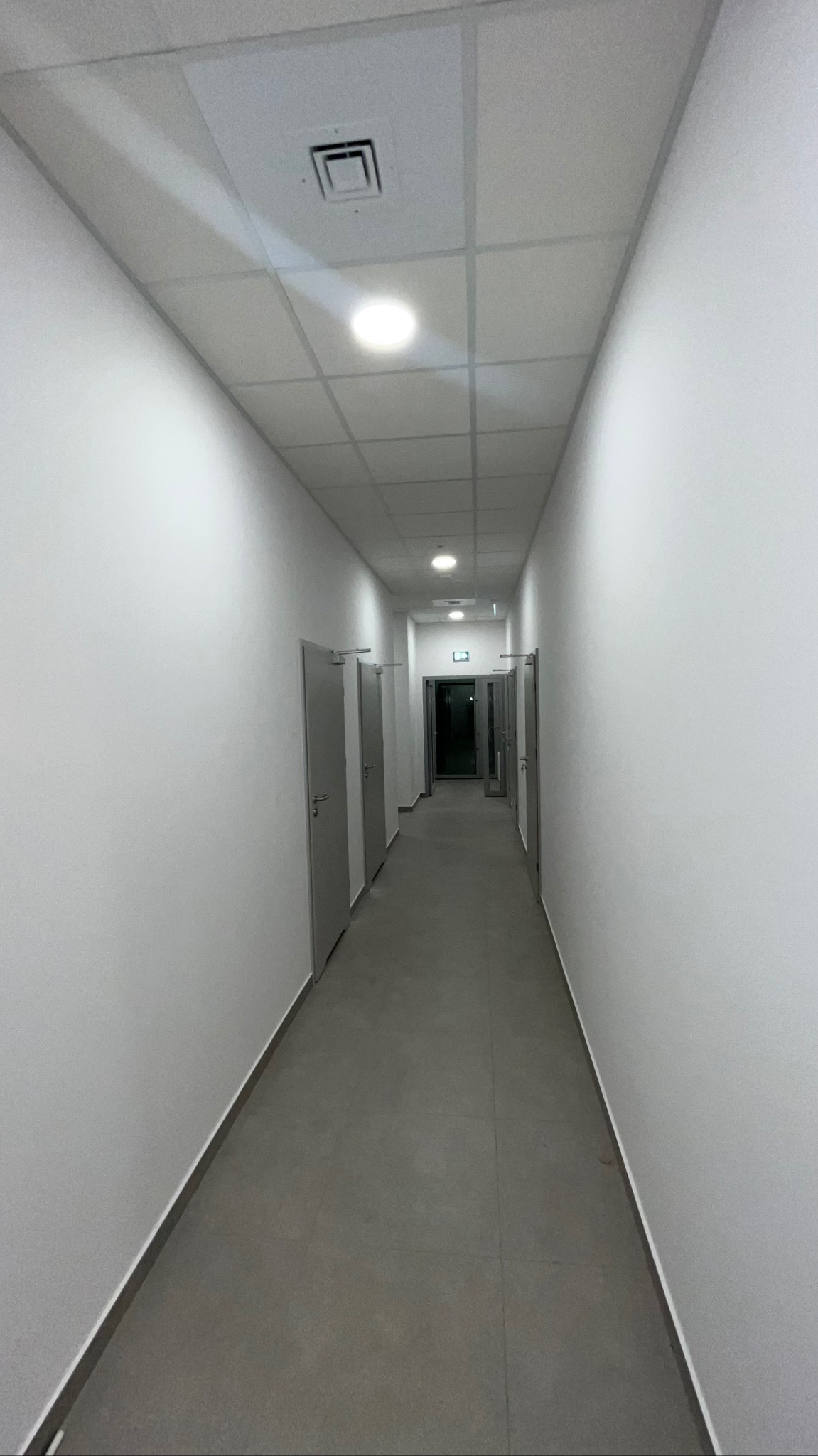 Długi, pusty korytarz z szarymi drzwiami i podłogą z płytek. Białe ściany i sufit z oświetleniem. Widok w perspektywie, z wyjściem ewakuacyjnym na końcu.
