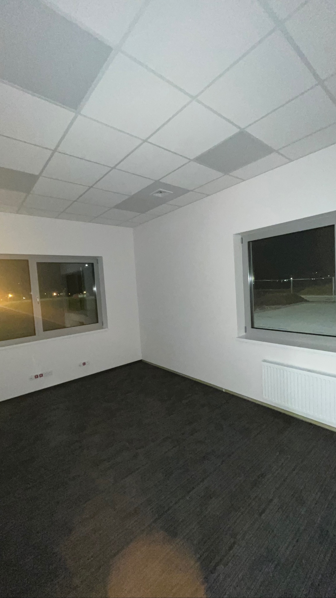 Pusty pokój biurowy z dwoma oknami, grzejnikiem i podwieszanym sufitem kasetonowym. Widok nocny za oknem. Ciemna wykładzina podłogowa.
