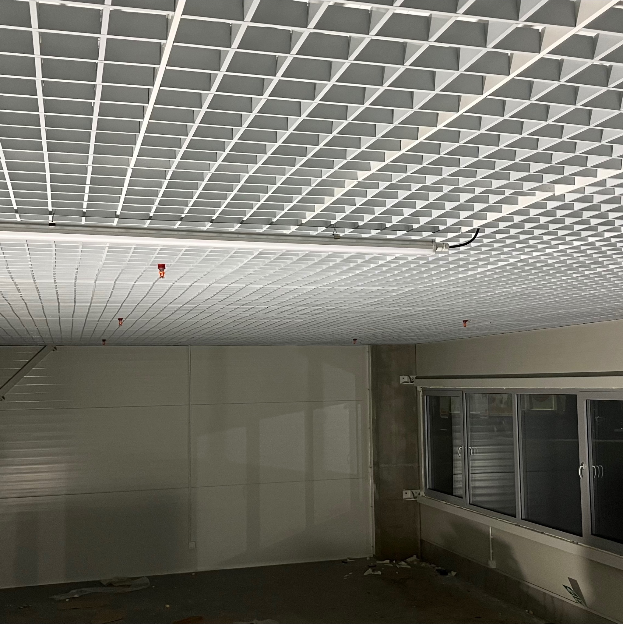 Biały sufit rastrowy z widocznymi instalacjami przeciwpożarowymi w pomieszczeniu z oknami. Widok z dołu, sufit w kratkę.