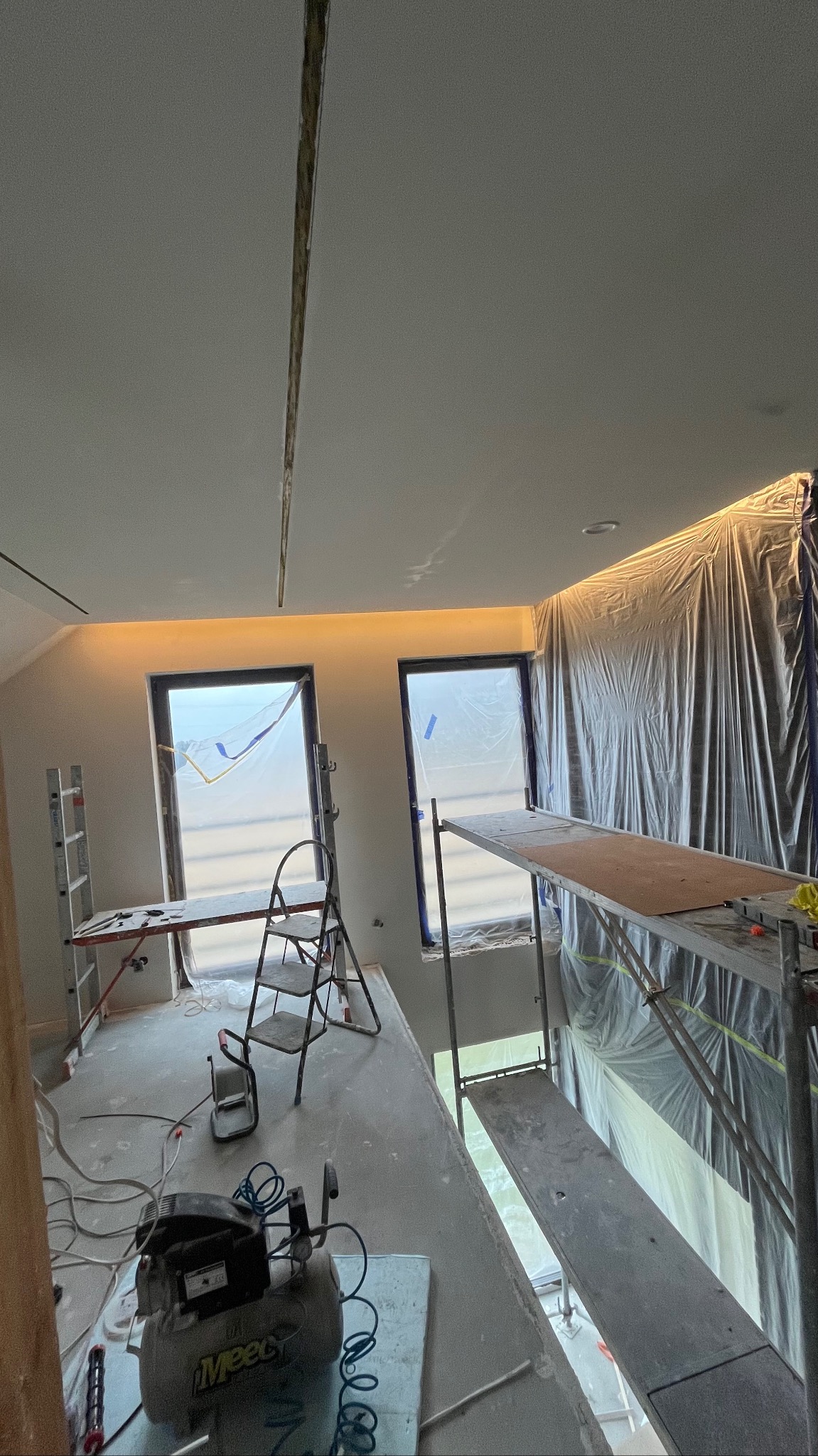 Wnętrze w trakcie prac wykończeniowych: rusztowania, okna zabezpieczone folią, widoczny kompresor. Przygotowanie do dalszych prac, oświetlenie w suficie.