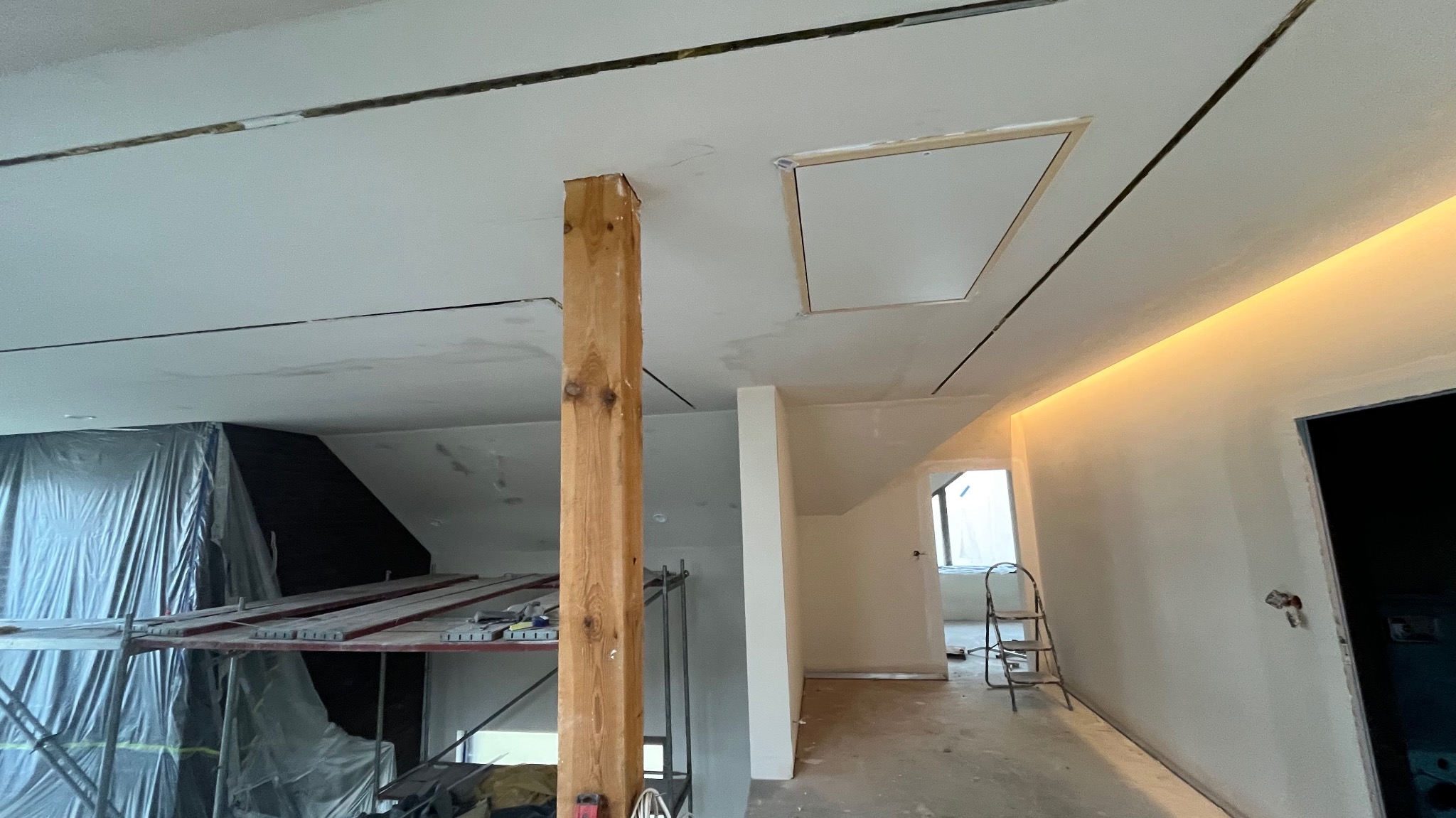 Sufit z płyt kartonowo-gipsowych w trakcie montażu, widoczne łączenia, drewniany słup podtrzymujący konstrukcję, rusztowanie i oświetlenie LED wzdłuż ściany.