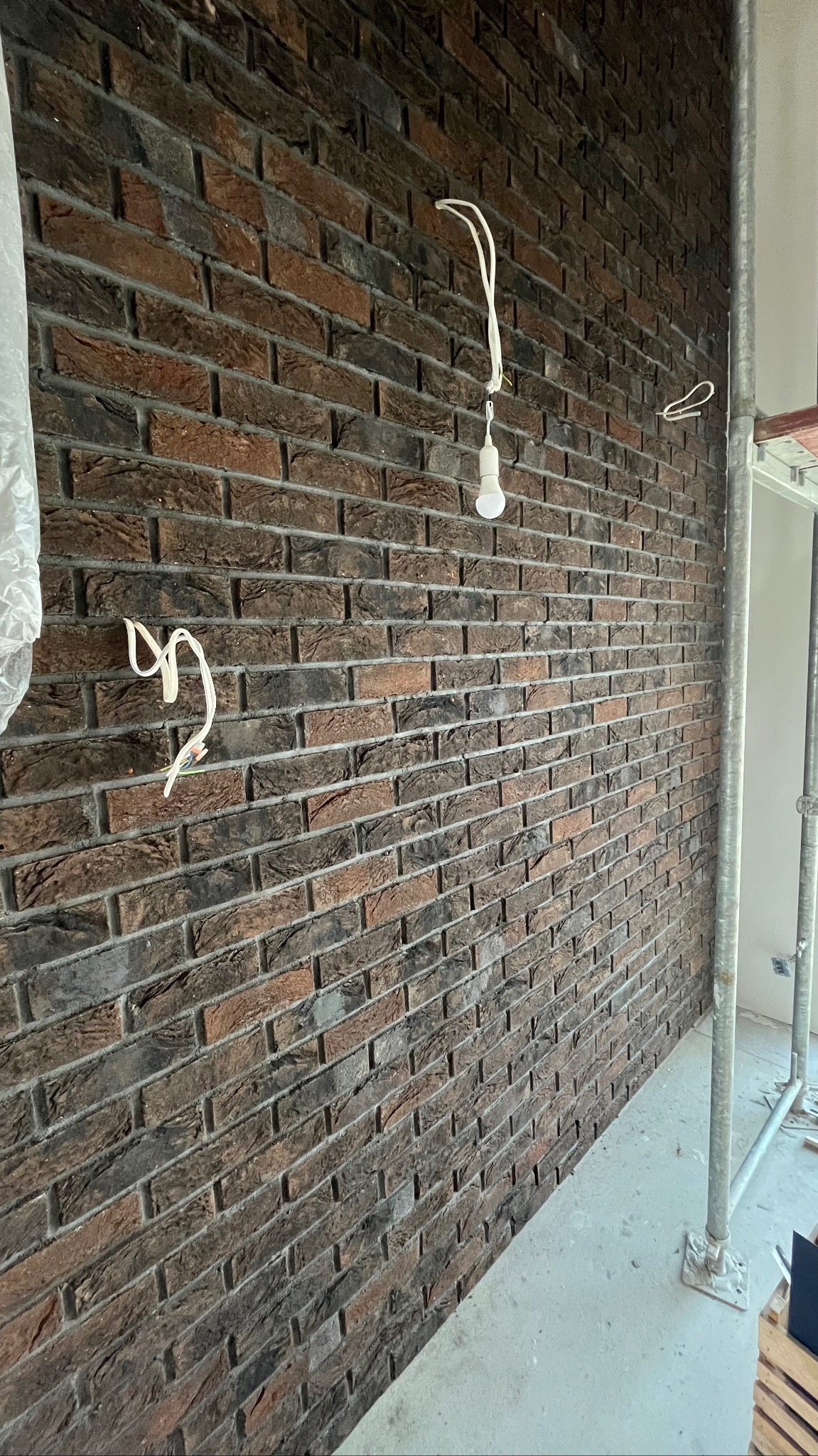 Ściana z cegły klinkierowej w odcieniach brązu i szarości, z widocznymi przewodami elektrycznymi i żarówką. Fragment rusztowania w tle. Układanie cegieł w trakcie prac wykończeniowych.