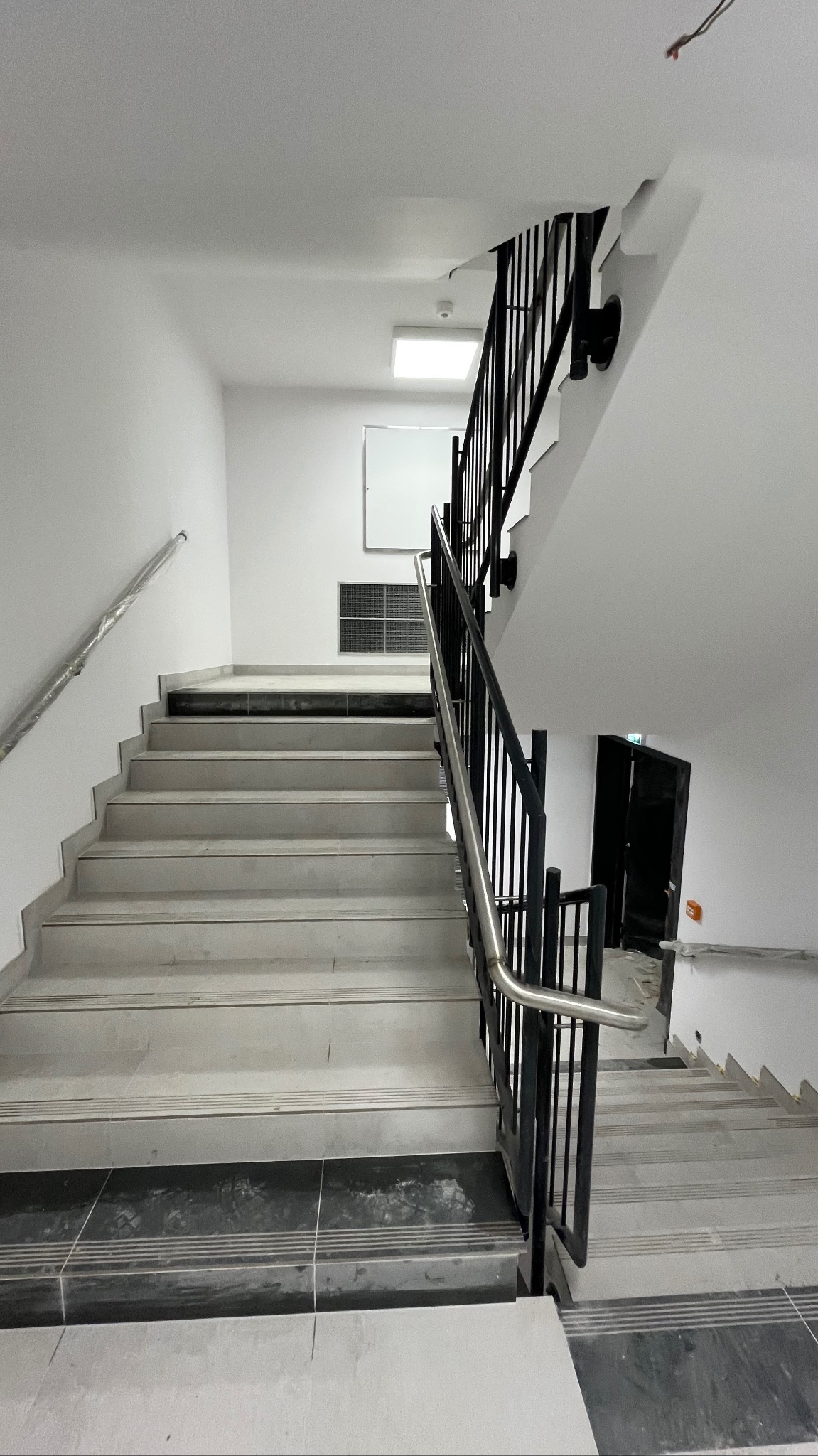 Klatka schodowa w jasnej tonacji z czarną metalową balustradą i szarymi płytkami na schodach. Widoczne wykończenia i detale architektoniczne. Minimalistyczny design.