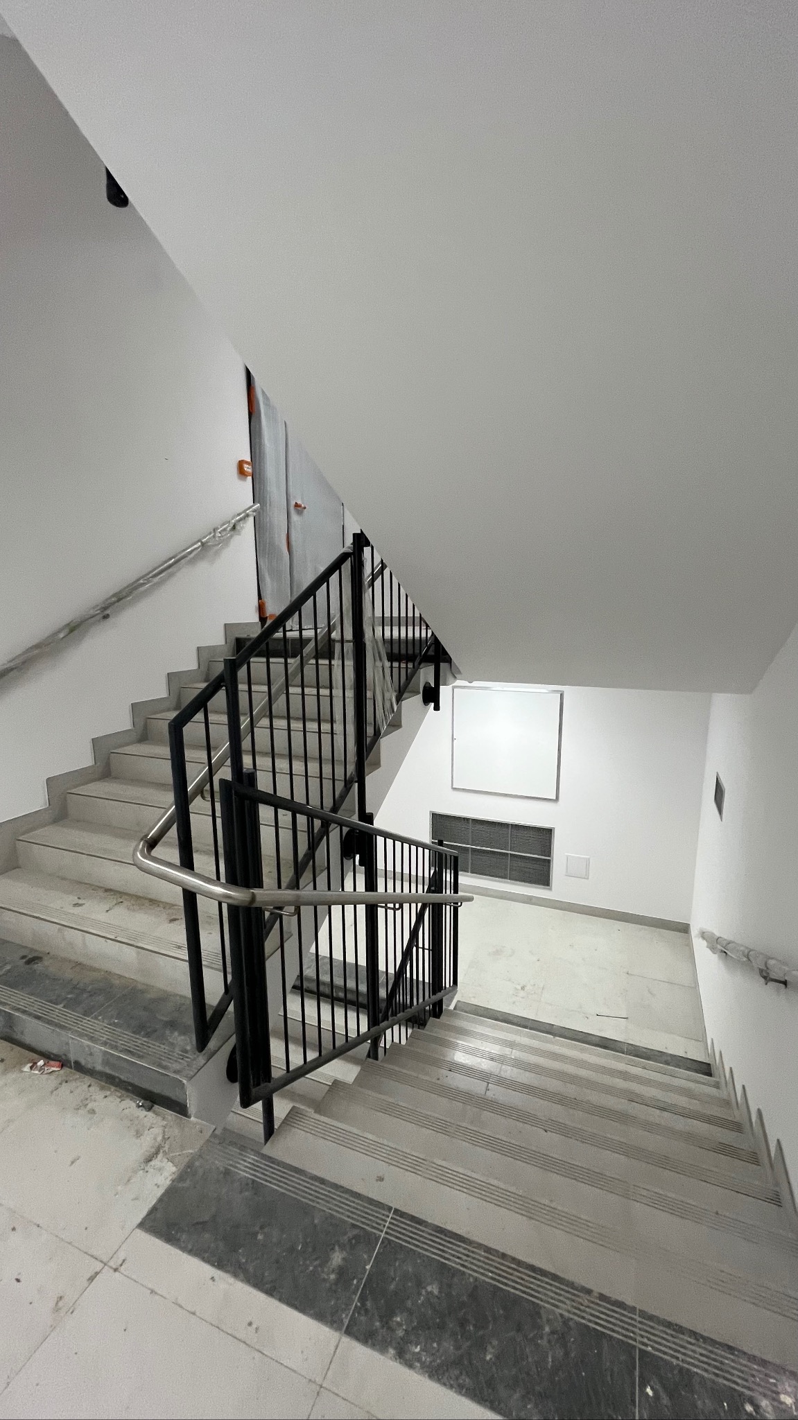 Klatka schodowa w trakcie wykańczania. Widoczne schody z szarymi płytkami, czarna metalowa balustrada i białe ściany. Ujęcie z góry, podkreślające perspektywę.