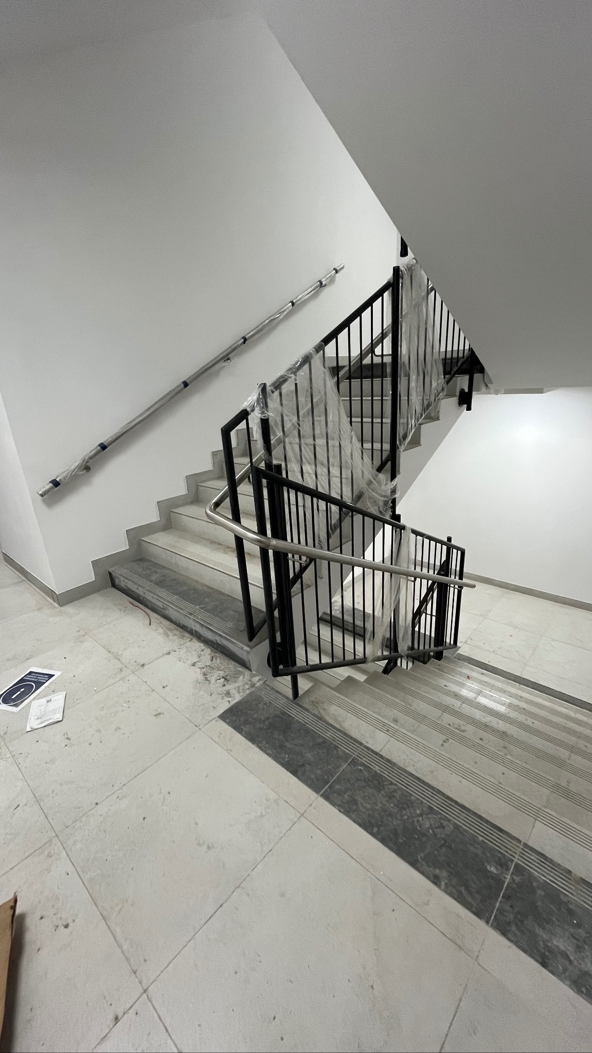 Klatka schodowa w trakcie wykańczania: białe ściany, schody z szarymi akcentami, czarne metalowe barierki częściowo zabezpieczone folią. Widoczne ślady po pracach.