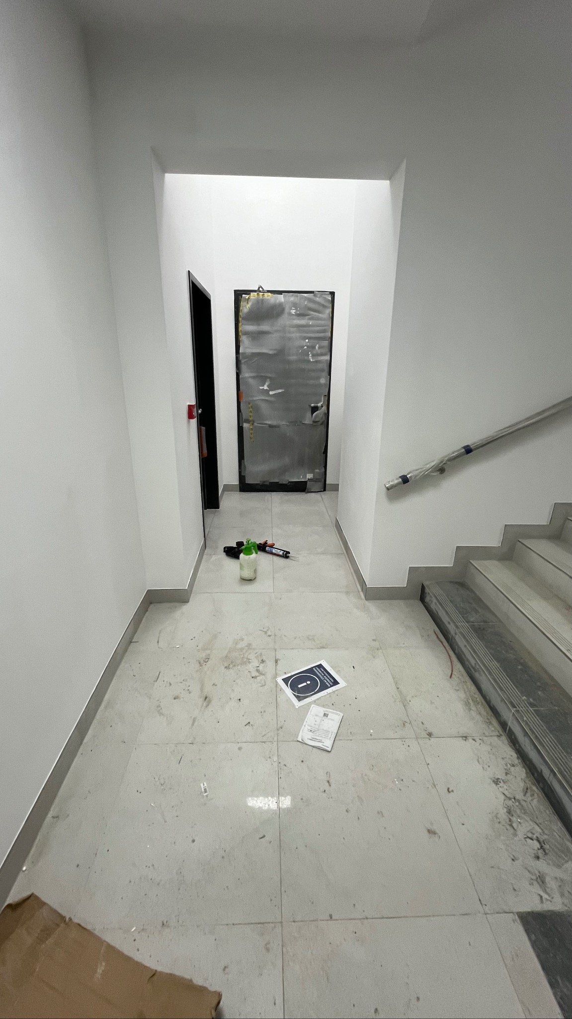 Korytarz w trakcie remontu: białe ściany, szare płytki podłogowe z zabrudzeniami, zabezpieczone drzwi, narzędzia i kartony na podłodze. Widok z dołu.