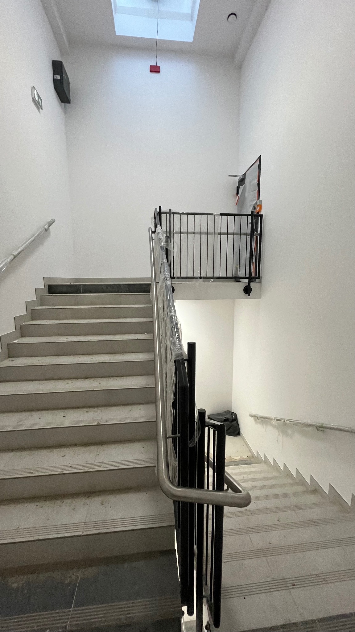 Klatka schodowa w trakcie wykańczania, zabezpieczone barierki, widoczne schody z szarej terakoty, białe ściany i sufit z oknem dachowym. Minimalistyczny styl.