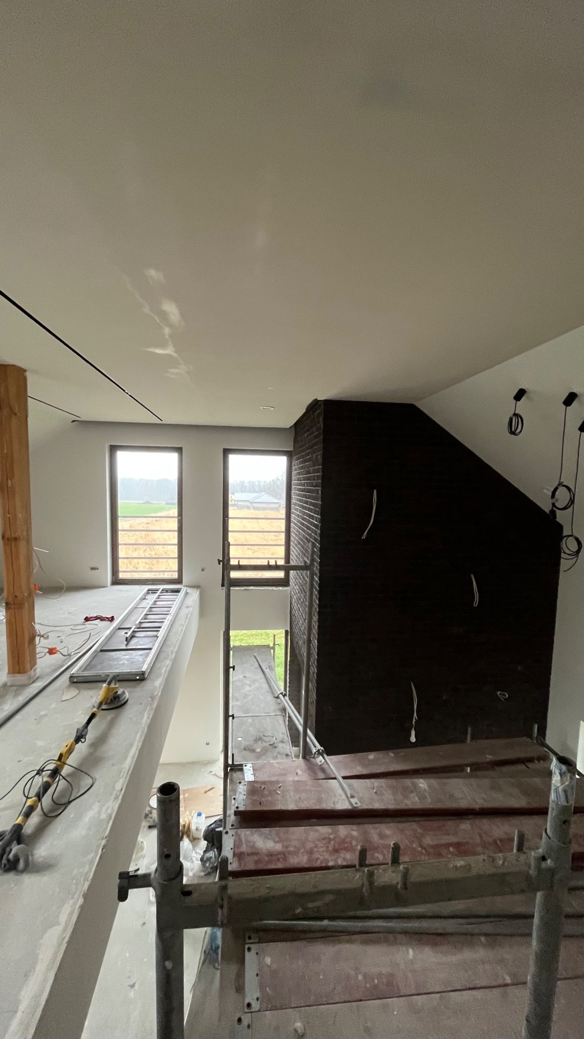 Wnętrze w trakcie prac wykończeniowych: widok na antresolę z rusztowaniem, fragment ściany z cegły klinkierowej, okna z widokiem na zewnątrz, narzędzia na posadzce.