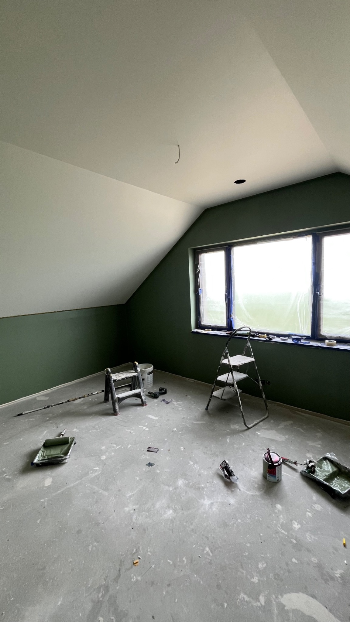 Pokój na poddaszu po malowaniu ścian na zielono, widoczne drabiny, kuwety malarskie i zabezpieczone okno folią. Ślady farby na podłodze.