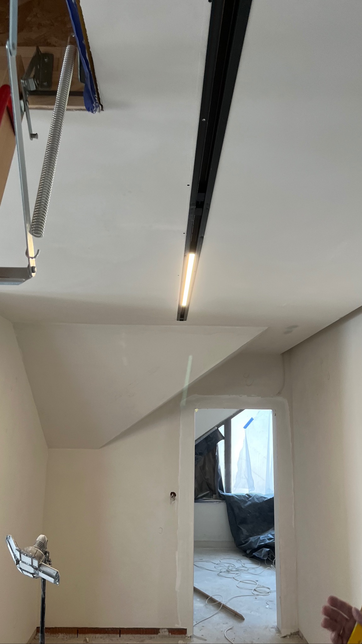Białe wnętrze w trakcie prac wykończeniowych: sufit z zamontowanym oświetleniem szynowym, ściany po szpachlowaniu, widoczne narzędzia i zabezpieczenia okienne.