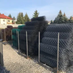 Skladowane rolki siatki ogrodzeniowej w różnych kolorach (czarny, zielony, brązowy) na żwirowym podłożu, z widocznymi metalowymi słupkami i budynkiem mieszkalnym w tle.