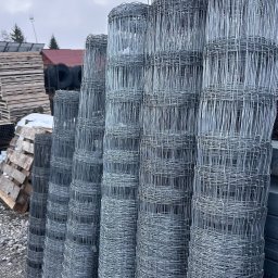 SIATKO-MET PAWEŁ ŚNIEŻEK - Stos rolek siatki ogrodzeniowej na żwirowym podłożu, widoczne palety i opony w tle, pochmurne niebo. Materiał do budowy ogrodzenia.