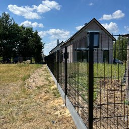 SIATKO-MET PAWEŁ ŚNIEŻEK - Nowoczesne ogrodzenie panelowe z podmurówką betonową, widok z perspektywy ścieżki wzdłuż ogrodzenia, dom w tle, błękitne niebo z chmurami.
