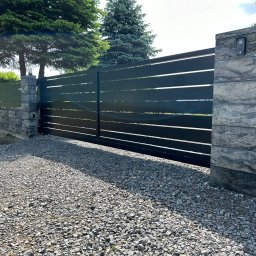 SIATKO-MET PAWEŁ ŚNIEŻEK - Nowoczesna, przesuwna brama z poziomych profili, zamontowana między kamiennymi słupkami z domofonem. Podłoże wysypane grysem, w tle drzewa.