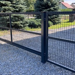 SIATKO-MET PAWEŁ ŚNIEŻEK - Grafitowa brama wjazdowa z siatki panelowej na żwirowym podłożu, z widocznym zamkiem i klamką, na tle zieleni i zabudowań.