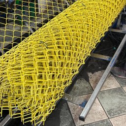 SIATKO-MET PAWEŁ ŚNIEŻEK - Żółta siatka ogrodzeniowa zwinięta w rolkę na metalowej konstrukcji maszyny produkcyjnej. Widoczne detale łączeń i faktura siatki.