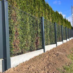 SIATKO-MET PAWEŁ ŚNIEŻEK - Nowoczesne ogrodzenie: szare panele z poziomymi szczelinami i siatka na betonowej podmurówce, tło z zielonych tuj, błękitne niebo. Widok z boku, lekki skos.