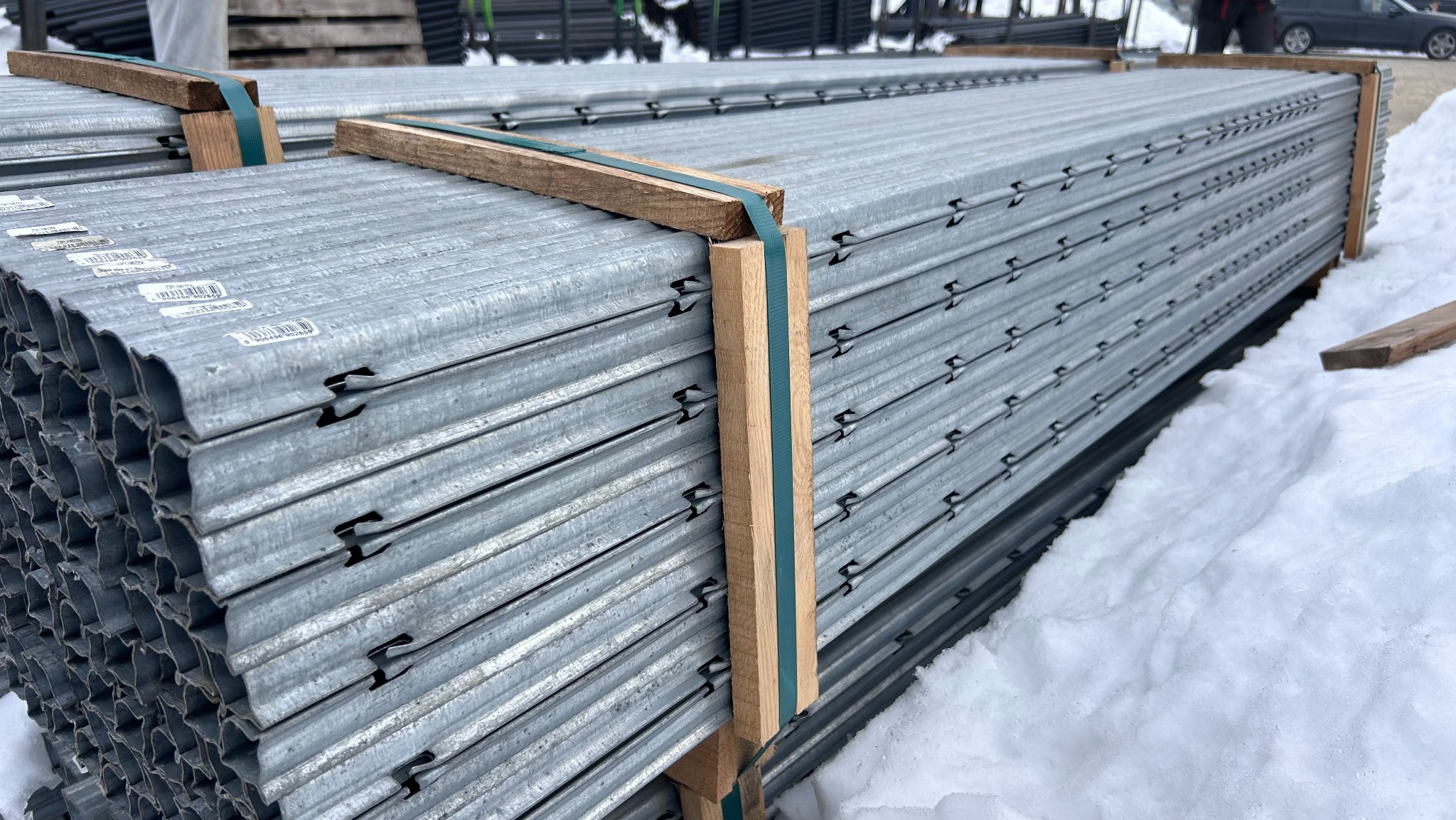 Stos ocynkowanych profili do montażu ogrodzenia panelowego, zabezpieczonych drewnianymi belkami i taśmą, leżący na śniegu. Widoczne zaczepy do paneli.
