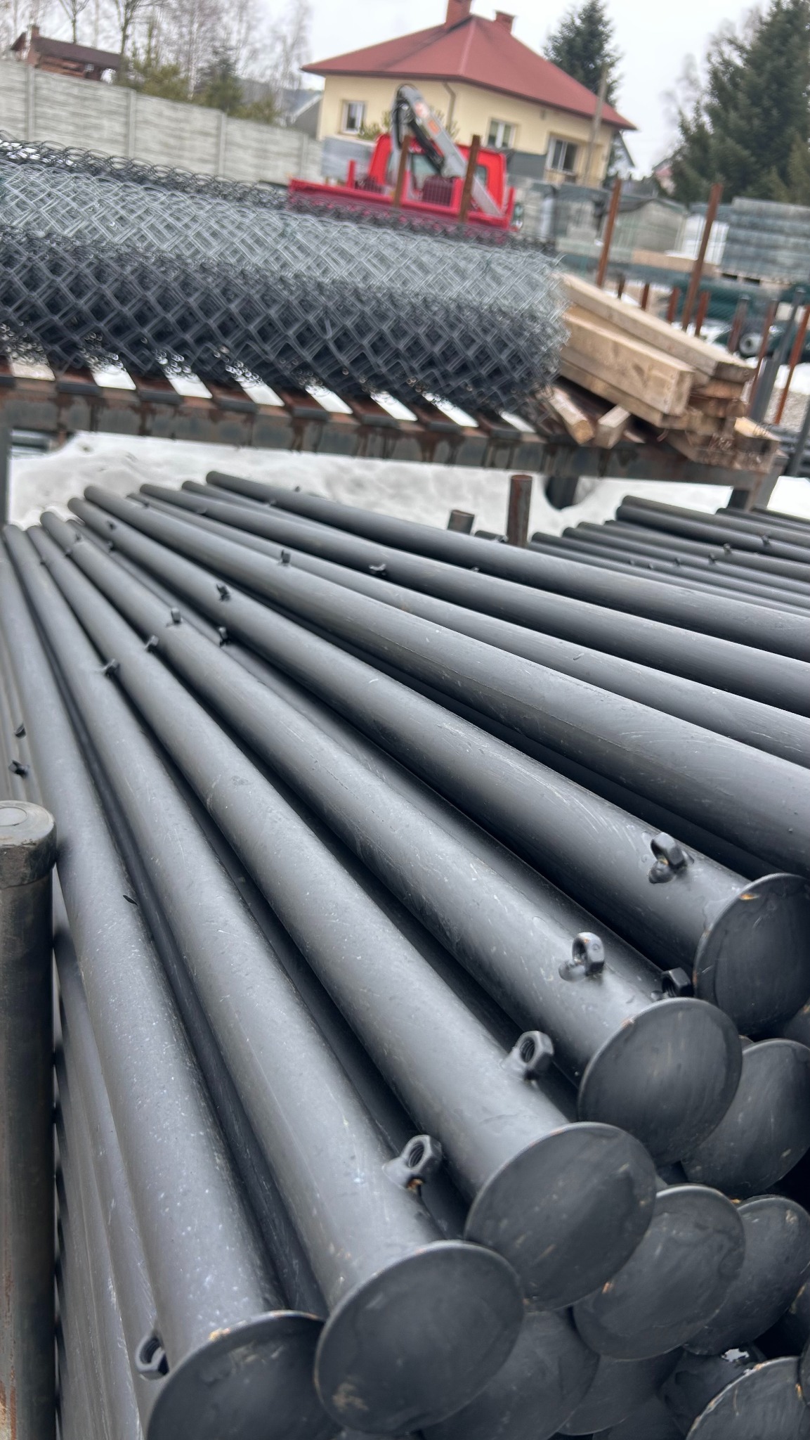 Stos czarnych, metalowych słupków z okrągłymi podstawami, w tle zwój siatki i budynek z czerwonym dachem. Elementy do montażu ogrodzenia.
