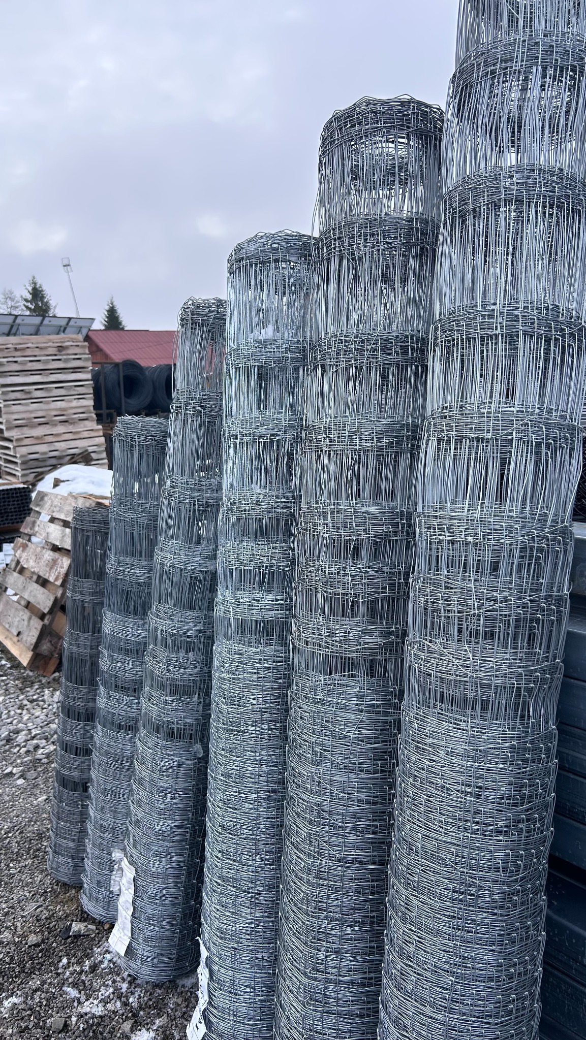 Stos rolek siatki ogrodzeniowej na żwirowym podłożu, widoczne palety i opony w tle, pochmurne niebo. Materiał do budowy ogrodzenia.